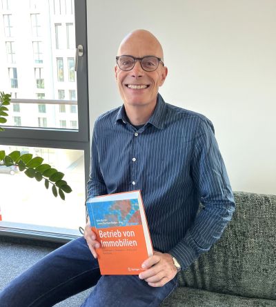 Andreas Klein sitzt auf der Couch mit dem Fachbuch "Betrieb von Immobilien" in der Hand